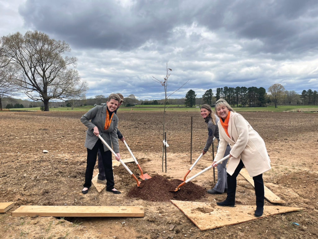 Four people with shovels smiling in front of a newly-planted tree