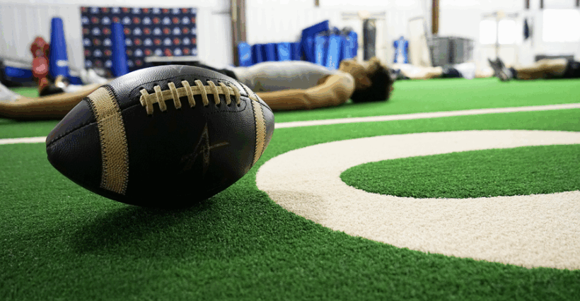 A football on an indoor practice field with players doing yoga in the background