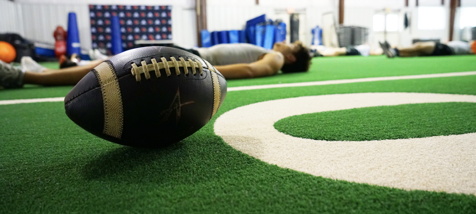 A football on an indoor practice field with players doing yoga in the background