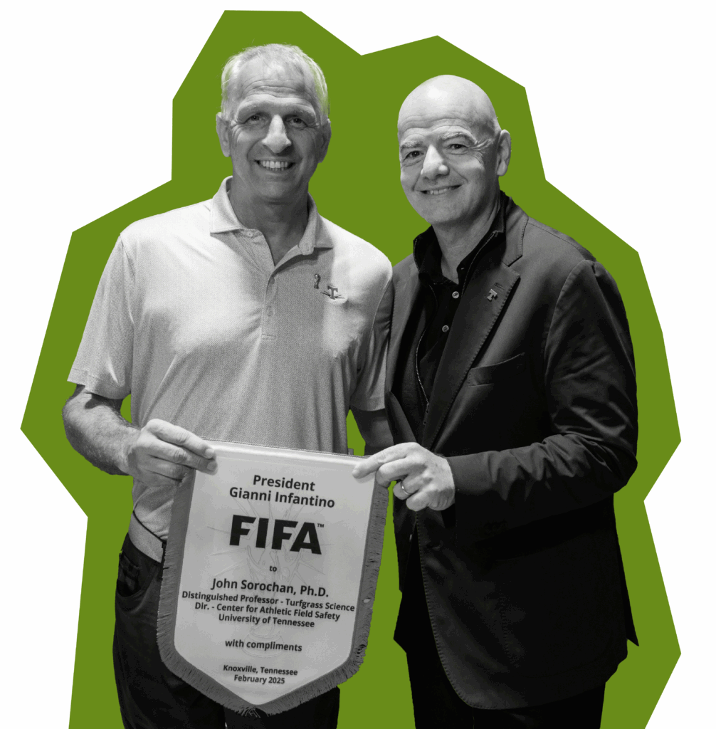 Two people smiling holding up a flag to honor John Sorochan that reads President Gianni Infantino FIFA to John Sorochan, Ph.D. Distinguished Professor - Turfgrass Science Director - Center for Athletic Field Safety University of Tennessee with compliments Knoxville, Tennessee February 2025