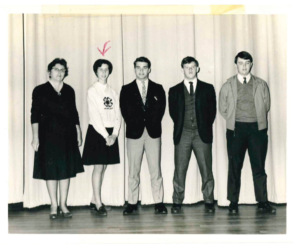 black and white photo of five people standing on a stage, including an arrow pointing to Ruth Henderson McQueen wearing a 4-H sweater