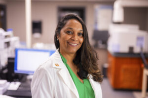 A smiling individual in a lab coat stands in a laboratory setting, wearing a green shirt and a white coat with a "T" pin, surrounded by scientific equipment.