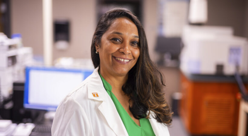 A smiling individual in a lab coat stands in a laboratory setting, wearing a green shirt and a white coat with a "T" pin, surrounded by scientific equipment.